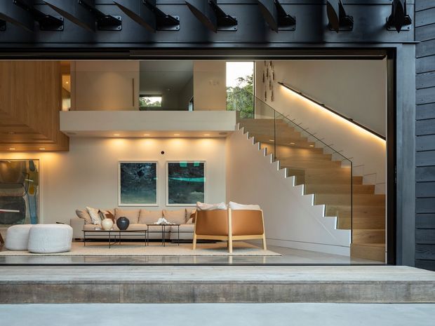 A contemporary residential interior viewed through a large open garage-style opening. The room features a warm, minimalist living area with a sofa, armchairs, coffee table, and pendant lighting, alongside a timber staircase with a clear glass balustrade and integrated warm LED lighting. Dark horizontal exterior cladding and exposed overhead garage-door hardware frame the view, while concrete thresholds and pale timber finishes add contrast.