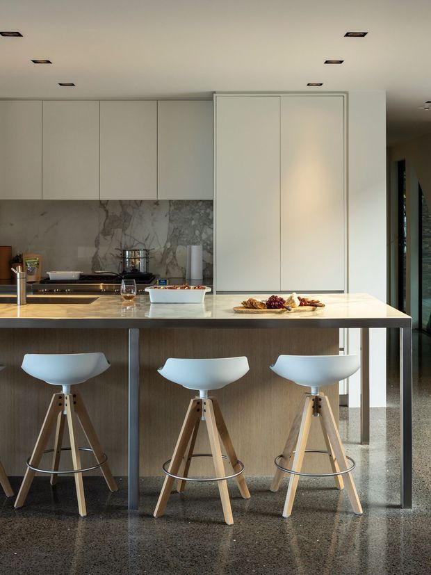 A refined modern kitchen with a long island and three white sculptural bar stools. The space features flat-panel white cabinetry, a large marble-look stone splashback, a pale stone benchtop, warm timber island joinery, integrated appliances, recessed ceiling lights, and polished grey terrazzo flooring.