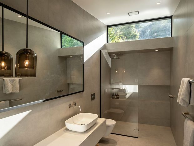 Contemporary bathroom with full-height warm grey tiled walls and floor, a floating pale vanity with a white vessel basin, wall-mounted tapware, toilet, and a frameless glass shower enclosure. A large black-framed mirror reflects pendant lights and towels, while high clerestory glazing brings in natural light and views of greenery. The space has a clean, refined, minimalist feel.