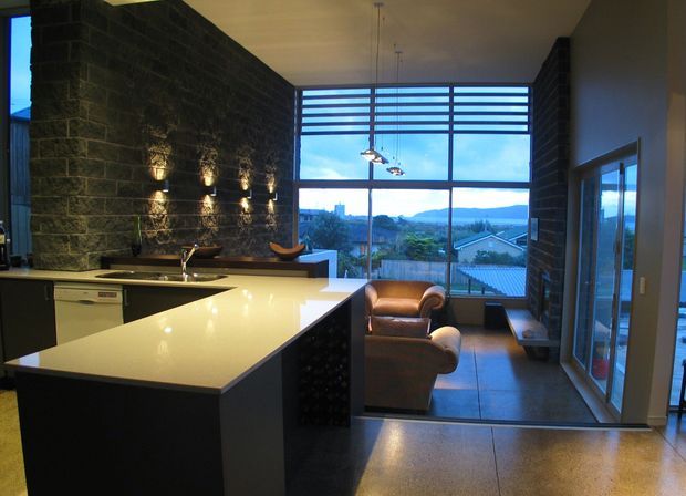 Open-plan contemporary kitchen and living area with a large white stone island, dark cabinetry, a dramatic dark masonry feature wall, polished concrete flooring, and full-height glazing overlooking a suburban landscape. Leather lounge chairs sit beside the window, with pendant lights suspended over the kitchen.