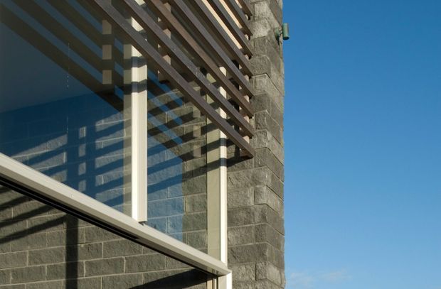 Close-up exterior architectural detail showing a dark grey block masonry wall, large reflective glazing, pale vertical structural posts, and a projecting timber or dark-stained horizontal louvre screen. Strong sunlight casts patterned shadows across the facade against a clear blue sky.