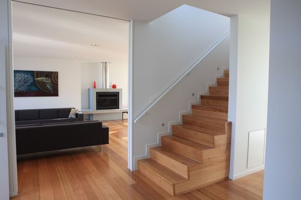 Interior view of a contemporary home showing a timber staircase with white stringers and balustrade, opening to a bright living room with dark sofas, a fireplace, artwork, and matching timber flooring. Clean white walls and recessed lighting create a minimalist, refined atmosphere.