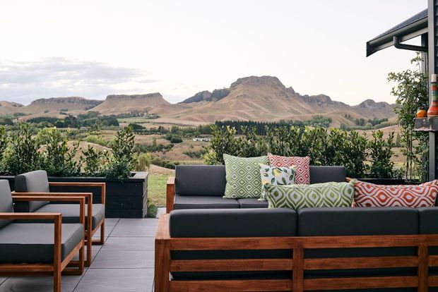 Outdoor covered terrace furnished with a substantial timber-framed lounge suite and matching armchairs, overlooking a broad rural valley and distinctive rugged mountain landscape. Large planters with greenery define the terrace edge, while patterned cushions add colour to the otherwise dark and natural-toned setting.