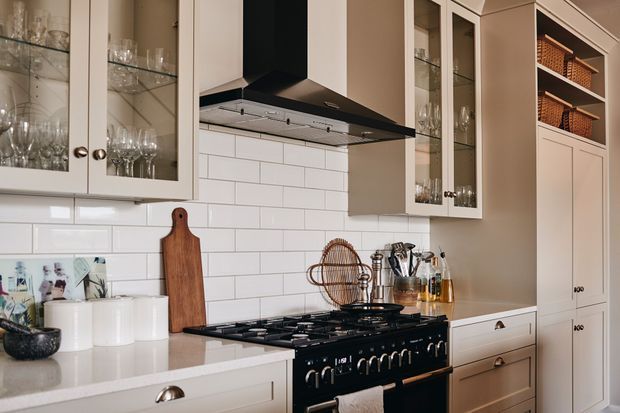 A bright, traditional-style kitchen with white shaker cabinetry, glass-front display cupboards, open shelving, a large black range cooker and hood, white subway-tile splashback, and pale stone benchtops. Glassware and cooking utensils are displayed throughout the cabinetry and work surfaces.