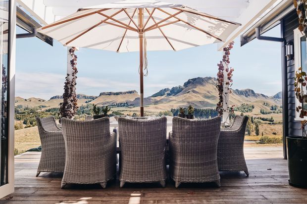 A covered outdoor dining terrace framed by open glazed doors, with a large cream umbrella over a dark woven dining table and chairs. Timber decking, climbing vines, and the surrounding dry mountain landscape are visible beyond.