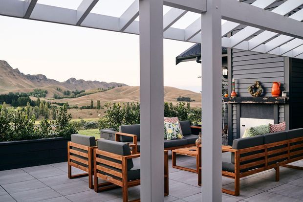 A covered outdoor terrace with a white pergola, grey tiled flooring, timber-framed lounge furniture, and dark grey built-in planters. A substantial outdoor fireplace with horizontal dark cladding forms the feature wall, while a dry mountainous rural landscape is visible beyond the terrace.