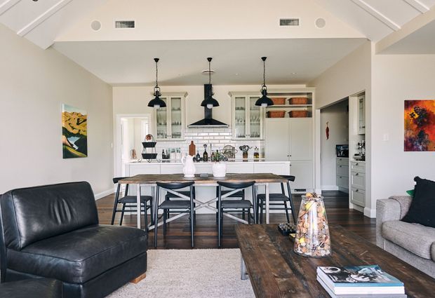 Open-plan interior with a dining area and kitchen featuring white shaker-style cabinetry, a large island with seating, pendant lights, dark timber flooring, and a vaulted white ceiling. A living area with leather and upholstered seating is visible in the foreground, creating a bright, relaxed contemporary home setting.