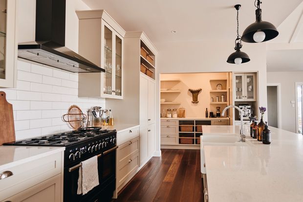 Bright, spacious kitchen with a large white island, classic white cabinetry, glass-front cupboards, open shelving, a black range cooker and prominent black pendant lights. The kitchen opens toward a butler’s pantry or additional storage area, with dark timber flooring and a white subway-tile splashback.