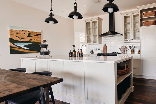A bright, traditional-style kitchen with a large white island, integrated open shelving, glass-front cabinetry, and a white subway-tile splashback. Black pendant lights and a black rangehood provide contrast, while dark timber flooring and a rustic timber dining table add warmth. A landscape artwork and decorative accessories complete the space.