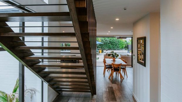 Contemporary open-plan interior viewed from a hallway beneath a dramatic open-riser timber staircase. The space features dark timber flooring, white textured walls, recessed ceiling lights, extensive glazing, and a dining area with timber furniture opening toward a leafy outdoor setting. A framed artwork is mounted on the right wall.