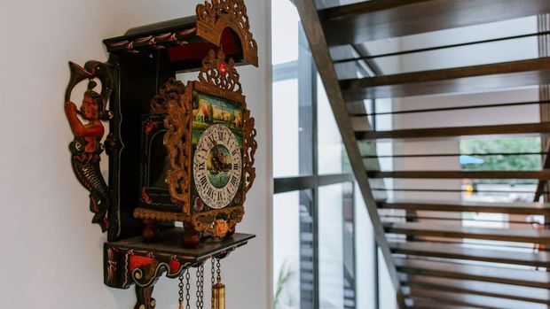 Interior architectural detail featuring an ornate dark wood carved wall-mounted clock beside a contemporary open-riser staircase with dark timber treads and slim metal supports. Glazing and a bright adjoining space are visible beyond the stair.