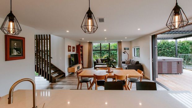 Open-plan contemporary living and dining area viewed from a kitchen island, with a pale stone-look benchtop in the foreground, timber dining table and chairs, lounge seating, a staircase with vertical timber screening, pendant lights, and large glazed openings connecting to a covered outdoor deck with a spa. The space has warm neutral finishes and abundant natural light.
