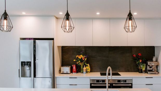 Contemporary kitchen with white full-height cabinetry, stainless-steel French-door refrigerator, dark stone-look splashback, integrated oven and cooktop, and a central island with a curved brushed-metal tap. Three geometric black pendant lights provide contrast.