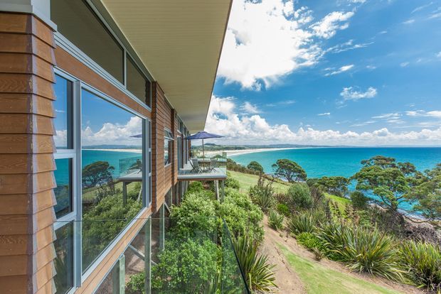 Exterior side view of a contemporary coastal home overlooking a bright turquoise bay. The building features horizontal timber cladding, large glazed windows, a broad projecting roof eave, and a small elevated balcony or deck. Native coastal planting and sandy terrain surround the house, with expansive ocean and beach views beyond.
