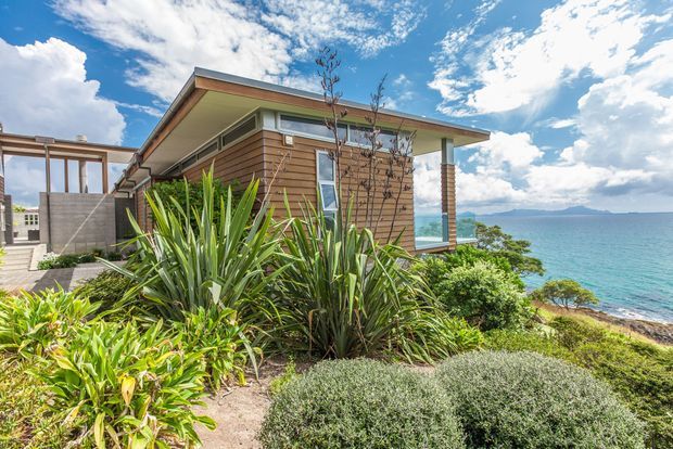 Exterior view of a low-slung contemporary coastal home set above a turquoise ocean. The building has horizontal timber cladding, broad roof overhangs, large glazing, and a covered outdoor connection, surrounded by dense coastal landscaping with grasses and shrubs.