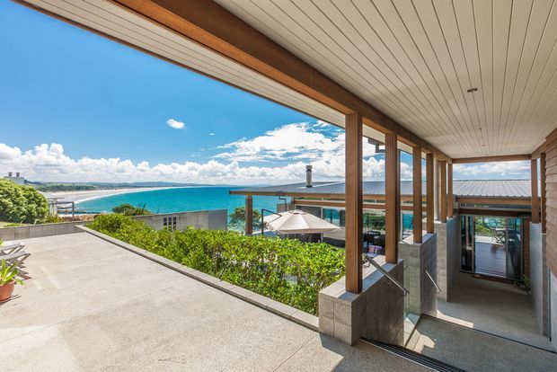 A bright covered outdoor terrace and walkway overlooking a vivid blue coastal bay. The space features a broad concrete-paved terrace, timber-framed posts and beams, a white-lined soffit ceiling, stone or concrete-clad column bases, and glazed openings leading toward the house. A pool, outdoor seating, hedging, and neighbouring roof forms are visible below, with expansive water views beyond.