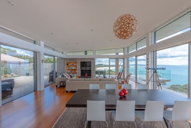 A bright, open-plan coastal living and dining space with extensive floor-to-ceiling glazing overlooking the ocean. Warm timber flooring extends through the room, with a large dark dining table and white chairs in the foreground, a contemporary lounge area and fireplace beyond, and a sculptural woven pendant light overhead. A telescope near the windows reinforces the strong seaside outlook.