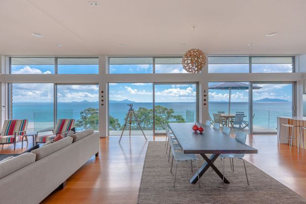 Bright open-plan waterfront interior with a living and dining area facing expansive floor-to-ceiling glazing. The room features warm timber flooring, a light upholstered sectional sofa, striped lounge chairs, a long dining table with modern crossed-leg base, a woven pendant light, and a small outdoor balcony or terrace beyond the glass overlooking turquoise water and distant mountains.