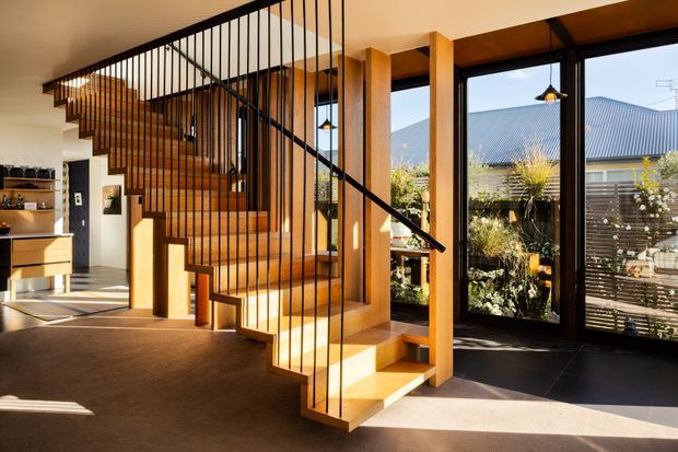 A bright contemporary interior centred on a sculptural timber staircase with open-riser steps and slender black metal balustrades. The stair connects an open living area to an upper level, with timber structural posts, warm timber cabinetry, dark flooring near the glazing, pendant lights, and large floor-to-ceiling windows overlooking a planted courtyard and neighbouring roofline.