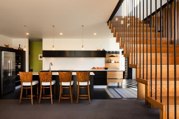 Open-plan contemporary kitchen and dining area with a long black island, integrated dark cabinetry, white splashback, stainless-steel refrigerator, timber bar stools, and pendant lighting. A striking timber staircase with slender black vertical balustrades dominates the right side, creating a strong architectural feature. The space has a warm, refined palette with timber accents and a charcoal floor.