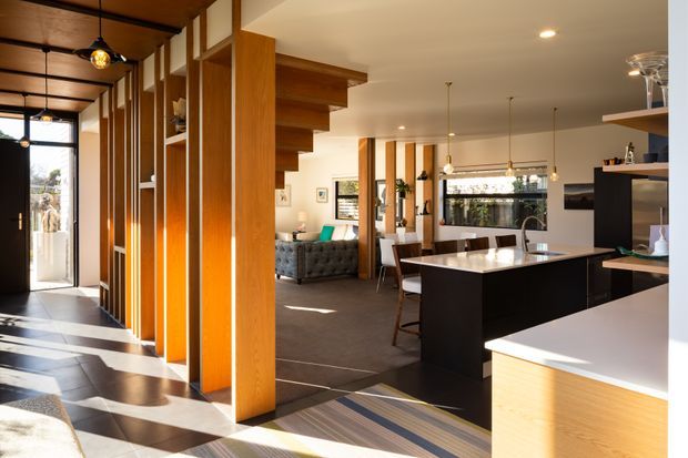 Open-plan contemporary home interior with a kitchen in the foreground, dining and living areas beyond, and a strongly expressed timber-framed entry and stair structure. Warm vertical timber battens and posts define the circulation zone, while a dark kitchen island with a light benchtop anchors the space. Large windows, pendant lights, recessed downlights, and patterned sunlight create a bright, refined interior.