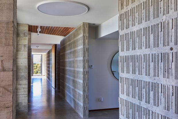 Interior view of a contemporary residential hallway featuring exposed textured concrete-block privacy screens, polished concrete flooring, timber slatted ceiling lining, and a large circular flush-mount ceiling light. The corridor opens toward bright glazing at the far end, with a curved mirror visible on the right.