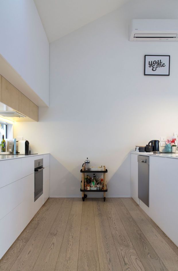 A bright, compact contemporary kitchen with white flat-panel cabinetry on both sides, light timber upper cabinets, pale timber flooring, and stainless-steel appliances. The room has a tall sloping ceiling, wall-mounted split-system air conditioner, and a small black metal drinks trolley centered against the rear wall. Minimal framed wall art and countertop appliances add simple decorative detail.