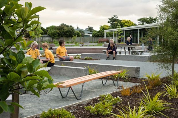A landscaped outdoor courtyard or communal garden beside a low modern building, featuring concrete seating terraces, timber benches, picnic tables, gravel paving, raised garden beds, and ornamental grasses. Several people are seated and socialising in the space. The setting has a contemporary, informal institutional or shared outdoor amenity character.