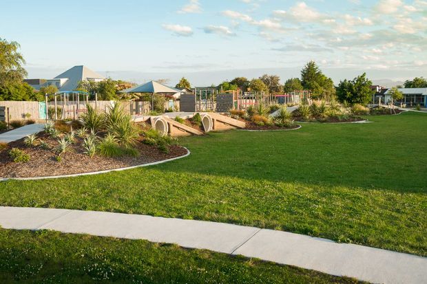 A landscaped outdoor recreation area with expansive manicured lawns, curved garden beds planted with grasses and succulents, concrete pedestrian paths, timber fencing, and small play or climbing structures. Low-rise buildings and trees sit in the background beneath a bright open sky.