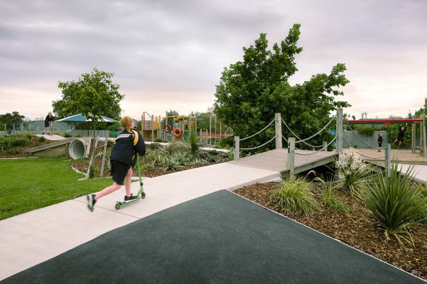A landscaped outdoor recreational space with smooth rubberized pathways, a small timber pedestrian bridge with rope handrails, garden beds of ornamental grasses and agave-like plants, lawn, and playground equipment in the background. A child is riding a scooter along the path beneath an overcast sky.