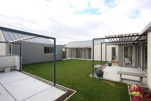 A contemporary single-storey courtyard-style residential development viewed from an outdoor paved area. The enclosed central lawn is surrounded by light-coloured rendered or panel-clad walls, dark metal-framed glazing, low-pitched metal roofs, and a timber pergola over a paved patio. Simple concrete paths, built-in outdoor seating, planters, and garden beds create a clean shared courtyard setting.