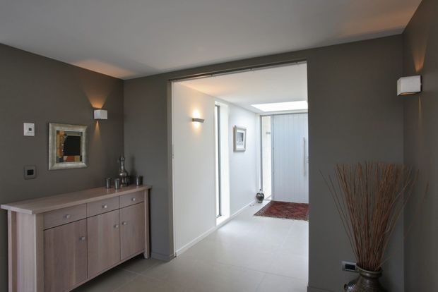 A contemporary residential entryway or hallway with dark taupe-grey walls, pale tiled flooring, a low timber sideboard, wall sconces, framed artwork, and a decorative vase of tall branches. The space opens into a bright white hallway with additional glazing and a red patterned rug.