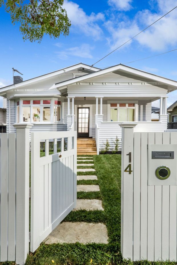Front exterior view of a single-storey white weatherboard house, photographed through an open picket gate. The home features a gabled roof, covered front porch, decorative timber posts and brackets, bay window, sash-style windows, front steps and a narrow grass-and-concrete path. A white fence and mailbox marked 14 frame the entry.