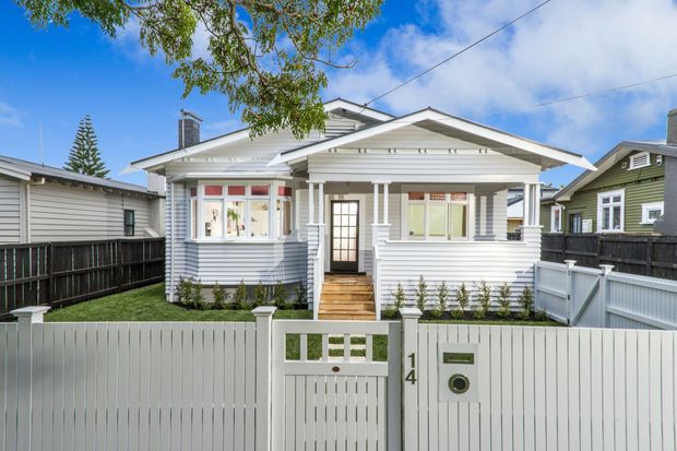 Front exterior view of a single-storey weatherboard bungalow with a prominent gabled verandah, white painted cladding, decorative timber trims, a small landscaped front yard, and a white picket-style boundary fence and gate.