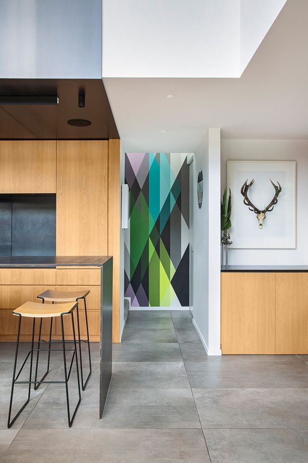 Contemporary open-plan interior showing a compact kitchen and adjoining entry or hallway. The kitchen features warm timber cabinetry, a dark stone-look benchtop, integrated appliances, a dark ceiling bulkhead, and two slim black-framed stools. Large grey floor tiles continue through the space. A vivid geometric multicolour artwork marks the hallway, while a framed antler motif and integrated timber storage add visual interest to the adjacent wall. The overall scheme is clean-lined with strong geometric accents and natural timber tones.