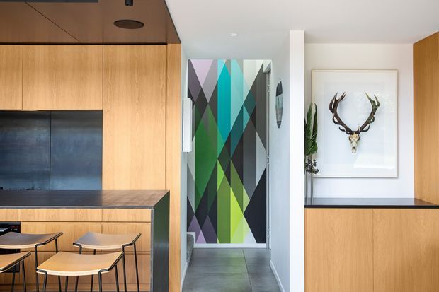 Bright contemporary kitchen and entryway featuring extensive warm timber cabinetry, dark benchtops, a compact island with stools, and a striking geometric multicolour feature panel on the hallway door. The white walls, grey floor, black accents, indoor plant, and framed sculptural antler artwork create a clean but bold interior.