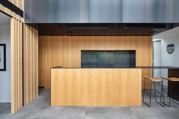 Contemporary kitchen with a large light-wood island and matching full-height wall cabinetry, dark overhead cabinetry, a dark stone or glass splashback, and a compact breakfast bar with stools. Vertical timber battens frame the left side, while large grey floor tiles and restrained black detailing create a clean, refined interior.