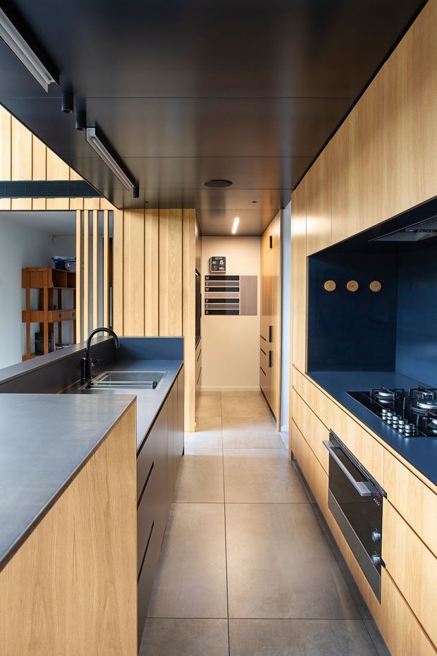 A narrow, galley-style contemporary kitchen with full-height light timber cabinetry on both sides, a large island with an integrated sink and dark blue-grey benchtop, and a matching dark blue splashback and cooking zone. The kitchen opens toward a hallway and adjacent space through a black-framed glazed partition with vertical timber battens. Large-format grey floor tiles and a dark metal ceiling create a refined, high-contrast interior, with integrated appliances, gas cooktop, concealed rangehood, and linear lighting.