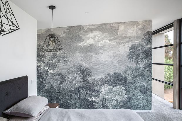 A contemporary bedroom with a low-profile upholstered bed, pale bedding, and a dramatic monochrome forest mural covering the feature wall. Black geometric pendant lights hang from the white ceiling, while a large black-framed glazed opening on the right brings in greenery and natural light. The room has a restrained grey, white, and black palette with a calm yet graphic character.
