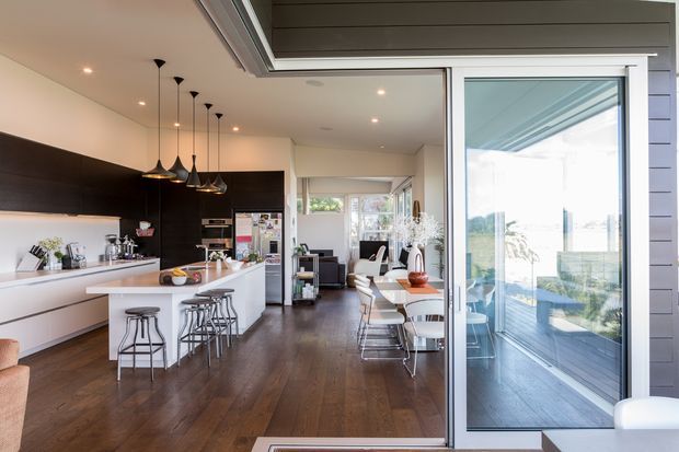 Bright open-plan contemporary kitchen and dining area with dark timber flooring, white cabinetry, a long island with bar stools, pendant lighting, and large glazed sliding doors opening to an outdoor deck. A living area is visible beyond the dining space.