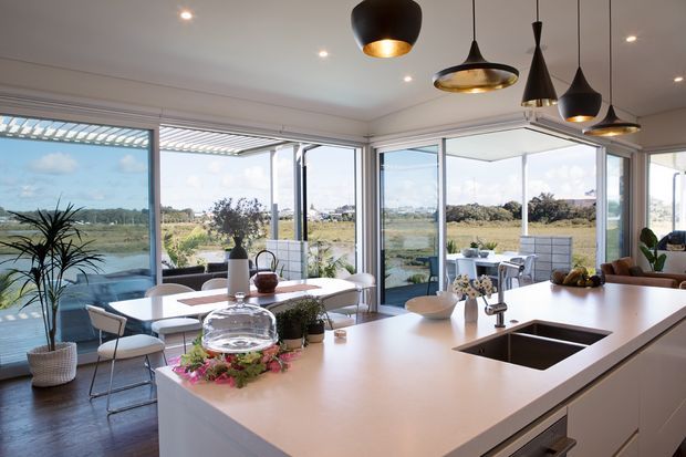 Bright, open-plan contemporary kitchen and dining area with a large pale benchtop island, integrated sink, white cabinetry, and a row of sculptural pendant lights. Full-height glazed sliding or folding doors open the room to a landscaped waterside outlook, while a dining table and indoor plants sit beside the glazing.
