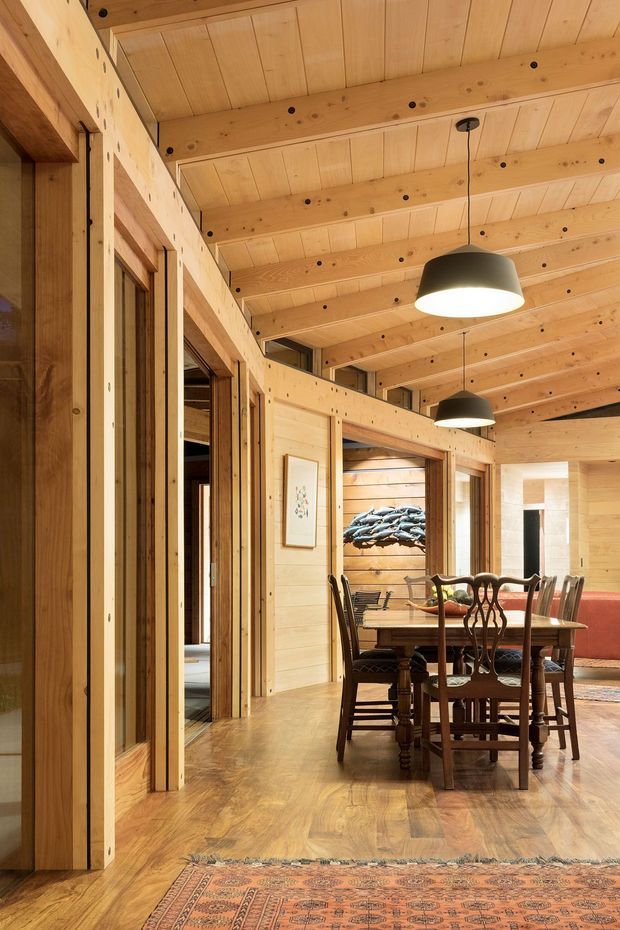 Warm timber-lined dining area in an open-plan lodge-style interior. The room features extensive light wood wall and ceiling panelling, exposed timber beams, timber flooring, dark pendant lights, and a traditional wooden dining table with upholstered and carved chairs. Doorways connect to adjacent rooms, while a large patterned rug and artwork add decorative detail.