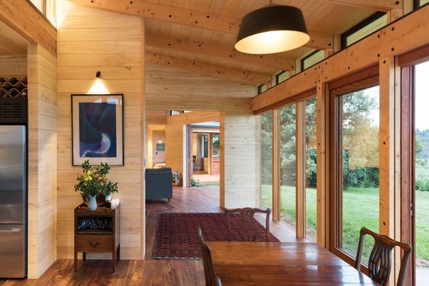 Warm timber-lined interior with an open-plan dining area in the foreground, featuring a large wooden dining table and chairs, floor-to-ceiling glazed doors and windows, a timber ceiling, exposed structural beams, and views to a leafy garden. A living area and hallway are visible beyond, with built-in wine storage and decorative furnishings along the left wall.
