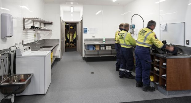 A bright, functional commercial washroom or utility preparation area with white tiled walls, grey seamless flooring, stainless-steel sinks, white cabinetry and shelving, a central stainless-steel workbench, and a wood-toned storage counter. Workers in high-visibility uniforms are using the counter and sink area. The space has clean, utilitarian finishes and overhead fluorescent lighting.