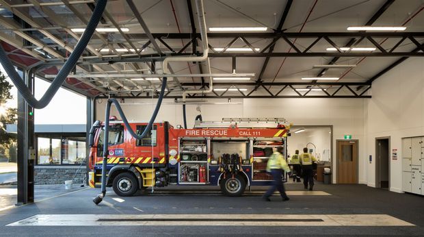 Interior view of a modern fire station apparatus bay, with a red fire rescue truck parked centrally, firefighters in high-visibility uniforms, polished concrete flooring, large glazed openings, exposed steel roof trusses, suspended linear lighting, and visible mechanical services.