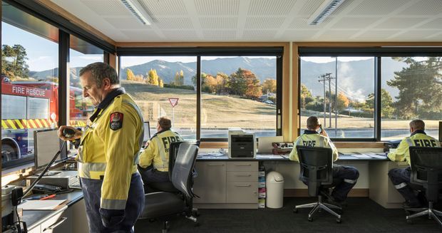 A bright operational fire and emergency services control room with several uniformed staff working at built-in desks and computer stations. Large floor-to-ceiling windows provide expansive views of a mountainous suburban landscape, while the interior features timber-framed glazing, light built-in cabinetry, dark carpet, and a perforated acoustic ceiling.