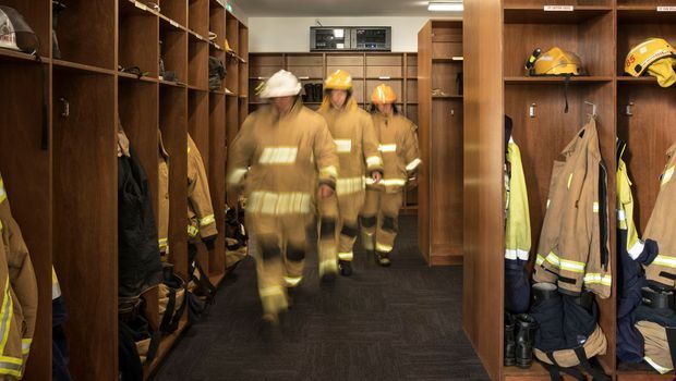 A locker room or changing area with dark timber open locker joinery lining both sides, firefighter helmets and protective uniforms stored in the compartments, and several firefighters walking through the space. The room has dark carpet flooring, warm artificial lighting, and a compact institutional fit-out.