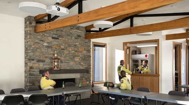 A bright, contemporary communal interior resembling a fire station or similar operational facility, with long meeting tables and chairs in the foreground, a large rough-stone fireplace wall, exposed timber beams and black steel framing overhead, circular ceiling lights, and a ceiling-mounted projector. A timber-fronted service or reception counter opens toward shelving and an adjacent room. People in high-visibility uniforms are present, but the architectural finishes and shared workspace dominate the image.