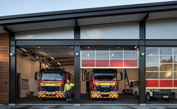 Exterior view of a contemporary fire station with three large vehicle bays. Two fire engines are parked in open bays, with a smaller vehicle visible in the right bay. The building has dark structural framing, broad glazed sectional bay doors with red horizontal panels, and a simple low-pitched roofline.