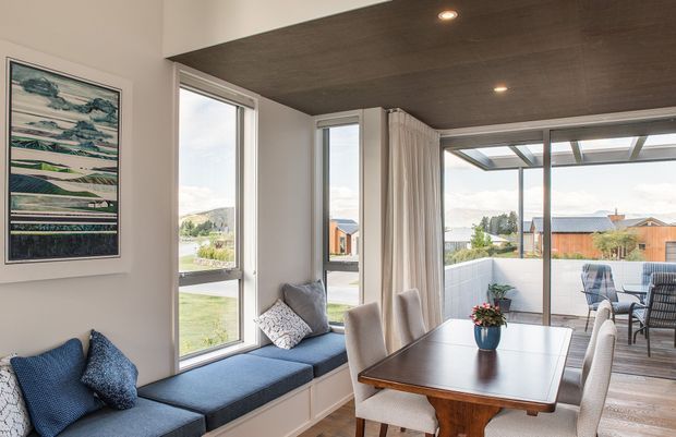 Bright contemporary dining and living space with a built-in window seat, blue upholstered cushions, a timber dining table, and upholstered dining chairs. Full-height corner windows and sliding glass doors open visually onto a covered outdoor deck and suburban landscape. The room features a dark timber-look ceiling, light neutral walls, timber flooring, and a framed abstract landscape artwork.